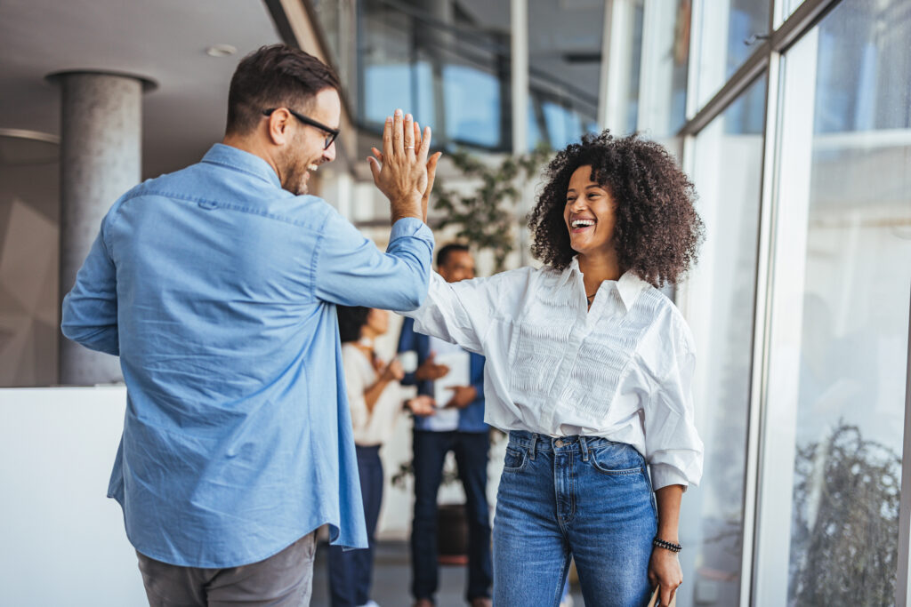Two happy business colleagues give a high five in a bright office, celebrating success and teamwork. They share joy and excitement, reflecting unity and a positive work environment.